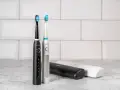 Dwa eleganckie modele szczoteczek sonicznych, czarny i srebrny, z różnymi końcówkami do wyboru. Obok etui podróżne.