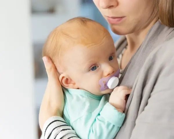 Malutkie dziecko z niebieskimi oczami i smoczkiem w buzi, tulone przez mamę. Zgryz od smoczka może być widoczny z czasem.